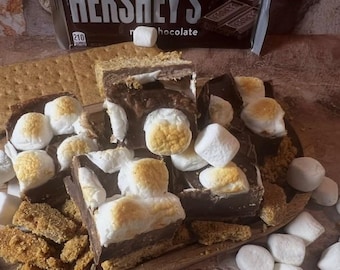 May include: Close-up of a s'mores dessert with graham crackers, chocolate, and toasted marshmallows. The Hershey's chocolate bar is visible in the background. The marshmallows are white with golden-brown tops. The dessert is on a wooden surface.