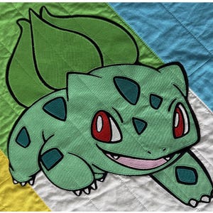Pode incluir: Aplicação de Bulbasaur verde sobre um fundo acolchoado com listras diagonais verdes, azuis, brancas e amarelas. O Bulbasaur tem olhos vermelhos, manchas verde-azulado e uma boca sorridente. A aplicação é delineada em preto.