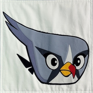 May include: A square white fabric panel featuring a large, stylized appliqué of a grey bird character with a pointed crest. The character has large white eyes with black pupils, a yellow beak with a red tip, and dark grey accents around the eyes.