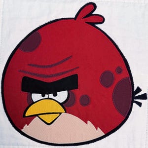 Może przedstawiać: Okrągła aplikacja z tkaniny przedstawiająca czerwoną postać z Angry Birds z jaskrawoczerwonym korpusem, grubymi czarnymi brwiami, białymi oczami, żółtym dziobem i jasnobeżową plamą na brzuchu, obszyta czarnymi nićmi na białym tle.