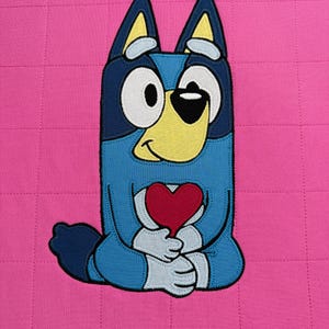 Può includere: Un personaggio di un cane blu dei cartoni animati che tiene un cuore rosso, su uno sfondo rosa trapuntato. Il cane ha grandi occhi, un muso giallo e orecchie blu. Il design è un'illustrazione giocosa e colorata.