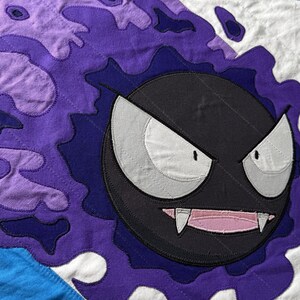 Puede incluir: Un edredón hecho a mano con un personaje de fantasma de dibujos animados con un cuerpo negro, ojos blancos y una boca rosa con colmillos. El edredón tiene una combinación de colores púrpura y azul, con un borde blanco.