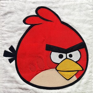 May include: A square white fabric panel featuring a large, red, circular bird appliqué with black eyebrows, white eyes with black pupils, a yellow triangular beak, and a light tan belly patch, all outlined in thick black stitching.