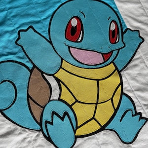 May include: A blue, yellow, and brown applique of a cartoon turtle character with a smiling face, red eyes, and a pink tongue. The turtle is on a white and blue quilted background. The character has a yellow shell and blue limbs.