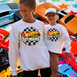 May include: A woman and child wearing matching white sweatshirts. The sweatshirts have a circular graphic with the text "Hot Mess Boymama" and "Mama's Boy" respectively. The background includes toy cars in various colors.
