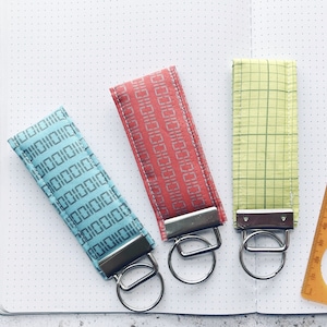 May include: Three keychains with different patterned fabric and silver key rings. The keychains are on a white notebook with a dotted grid pattern.