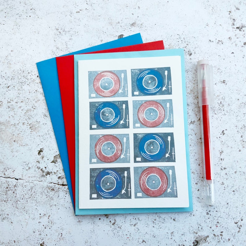 May include: A white card with a blue and red design of nine vinyl records on a turntable. The card is surrounded by three layers of paper in red, blue, and light blue.
