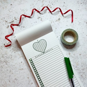 May include: A white notepad with a green gridded heart that says "I ♥ Spreadsheets". The notepad has a green pen and a roll of green tape on a white background.