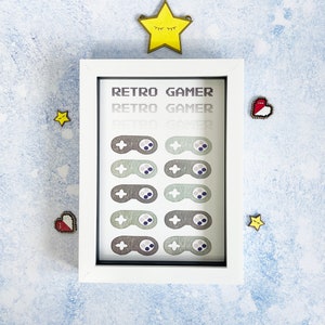 May include: White framed print with the text "Retro Gamer" repeated three times. The print features eight pixelated game controllers in shades of green and gray.