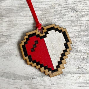 May include: A wooden heart-shaped ornament with a pixelated design. The heart is half red and half white, with a black outline.