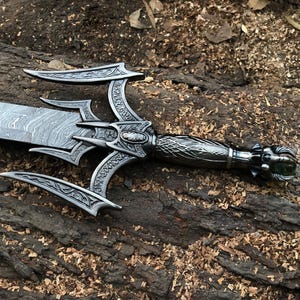 May include: A silver sword with a black, intricately carved handle and a green gem at the end. The blade is made of Damascus steel and has a wavy pattern.
