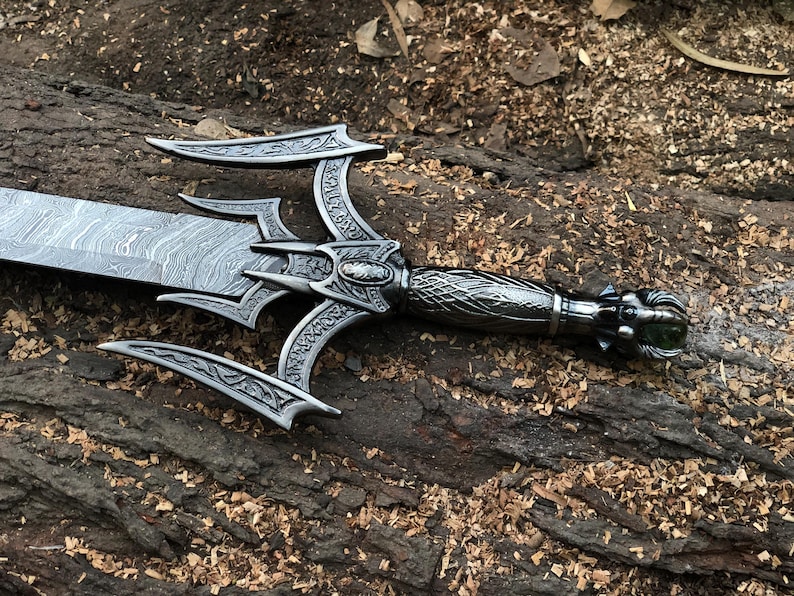 May include: A silver sword with a black, intricately carved handle and a green gem at the end. The blade is made of Damascus steel and has a wavy pattern.