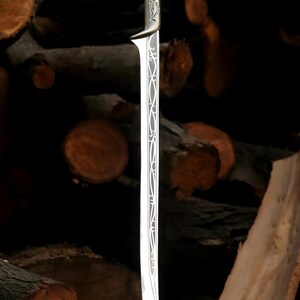 May include: A silver sword with an intricate design on the blade and handle. The sword is standing upright on a wooden surface.