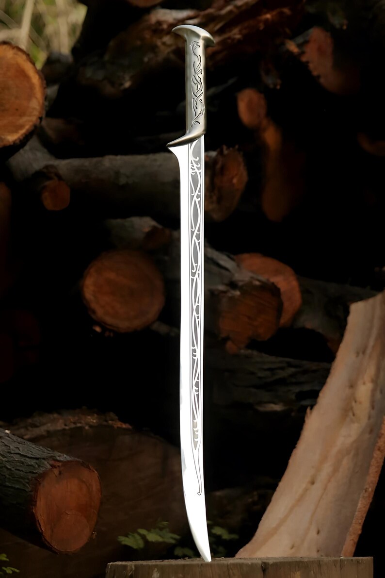 May include: A silver sword with an intricate design on the blade and handle. The sword is standing upright on a wooden surface.