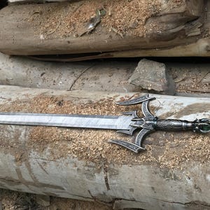 May include: A silver sword with a decorative handle and a green gem at the end. The sword is lying on a wooden surface.