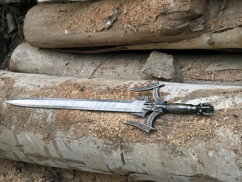 May include: A silver sword with a decorative handle and a green gem at the end. The sword is lying on a wooden surface.