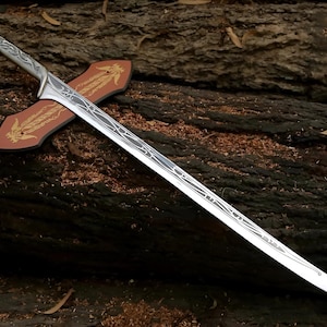 May include: A silver sword with a decorative handle and a wooden scabbard. The sword has a detailed design on the blade and is resting on a wooden surface.