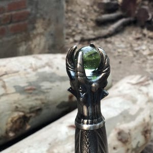 May include: Close-up of a sword hilt featuring an intricate dragon design. The hilt is topped with a green glass orb.