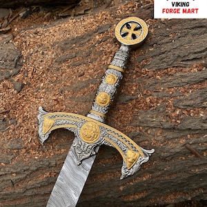 May include: An ornate sword with a silver and gold handle and crossguard, featuring intricate designs and a patterned blade. The sword rests on a wooden surface, with the "VIKING FORGE MART" logo in the upper right corner.