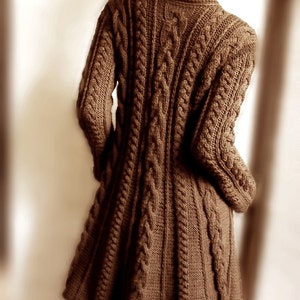 Könnte beinhalten: Ein brauner Strickpullover mit Zopfmuster und langer, fließender Silhouette. Der Pullover hat ein klassisches Zopfmuster und eine lockere Passform.