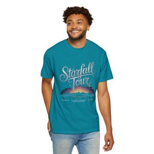 May include: A turquoise blue t-shirt with a graphic print of a mountain range with the text "Starfall Tour" in a stylized font. The text "Tour Locations" is printed below the graphic.