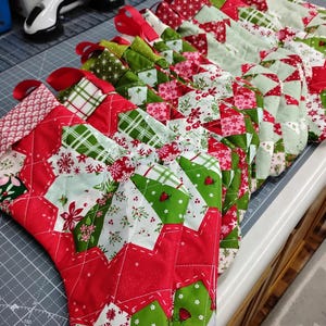 May include: A row of quilted Christmas stockings featuring a patchwork design in red, green, and white. The fabric patterns include snowflakes, plaid, and floral motifs. Each stocking has a red fabric loop at the top for hanging purposes.