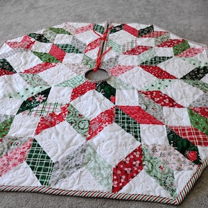 May include: A hexagonal quilted Christmas tree skirt featuring a patchwork star pattern in red, green, and white. The fabric displays various holiday-themed prints, including plaid, snowflakes, and floral motifs, with red ribbon ties at the center.