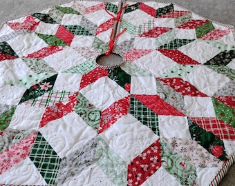 May include: A hexagonal quilted Christmas tree skirt featuring a patchwork star pattern in red, green, and white. The fabric displays various holiday-themed prints, including plaid, snowflakes, and floral motifs, with red ribbon ties at the center.