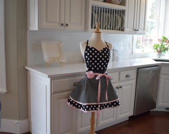 May include: A black and white polka dot apron with a pink ribbon and trim is displayed on a wooden mannequin. The apron is in a kitchen setting with white cabinets, a white countertop, and a stainless steel dishwasher.
