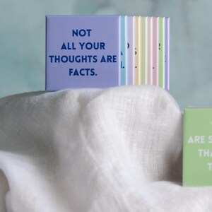 May include: A set of square magnets with pastel colors and motivational text. One lavender magnet displays the text NOT ALL YOUR THOUGHTS ARE FACTS in dark blue. A light green magnet displays the text YOU ARE STRONGER THAN YOU THINK in white.