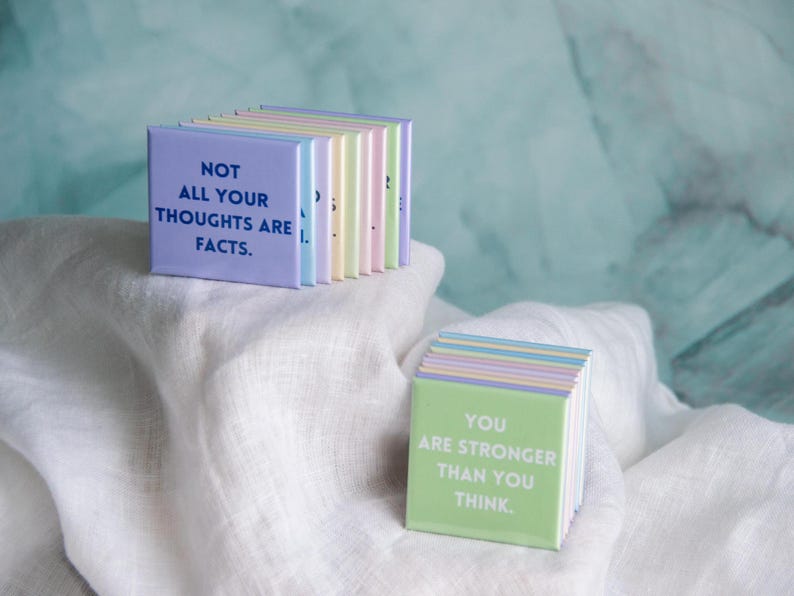 May include: A collection of square magnets in pastel colors. One stack features the text NOT ALL YOUR THOUGHTS ARE FACTS in dark blue. Another stack displays the text YOU ARE STRONGER THAN YOU THINK in white on a light green background.