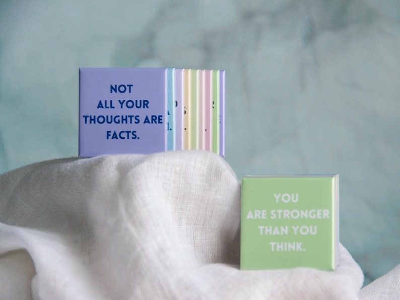 May include: A set of square magnets with pastel colors and motivational text. One lavender magnet displays the text NOT ALL YOUR THOUGHTS ARE FACTS in dark blue. A light green magnet displays the text YOU ARE STRONGER THAN YOU THINK in white.