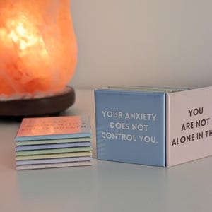 May include: A stack of small square magnets and two upright square magnets featuring text. The blue magnet reads, "YOUR ANXIETY DOES NOT CONTROL YOU." The pink magnet reads, "YOU ARE NOT ALONE IN THIS." The stack includes various pastel-colored magnets.