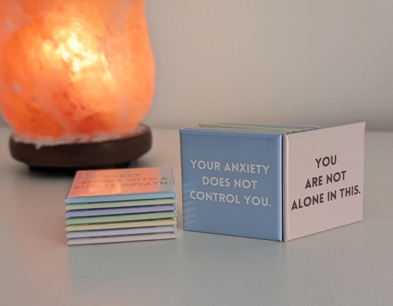 May include: A stack of small square magnets and two upright square magnets featuring text. The blue magnet reads, "YOUR ANXIETY DOES NOT CONTROL YOU." The pink magnet reads, "YOU ARE NOT ALONE IN THIS." The stack includes various pastel-colored magnets.