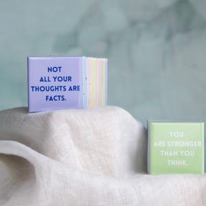 May include: A stack of square buttons in pastel colors, with the front button in light purple featuring the dark blue text NOT ALL YOUR THOUGHTS ARE FACTS. A separate light green square button sits nearby with the white text YOU ARE STRONGER THAN YOU THINK.