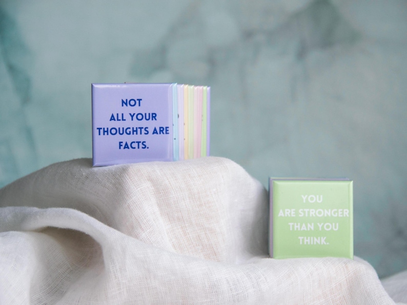 May include: A stack of square buttons in pastel colors, with the front button in light purple featuring the dark blue text NOT ALL YOUR THOUGHTS ARE FACTS. A separate light green square button sits nearby with the white text YOU ARE STRONGER THAN YOU THINK.