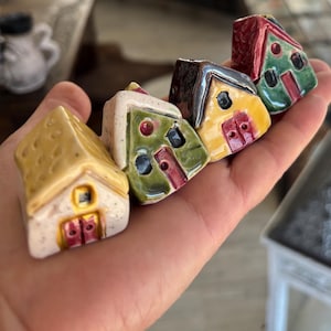 Può includere: Una mano mostra quattro casette in ceramica in miniatura. Ogni casa ha un design unico con colori diversi, tra cui giallo, verde, rosso e marrone. Le case presentano dettagli come porte, finestre e tetti strutturati, mostrando un'estetica stravagante e artigianale.