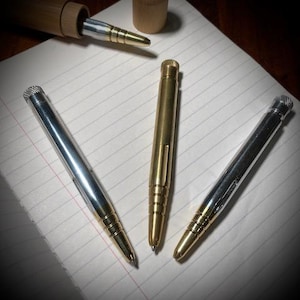 Könnte beinhalten: Drei Metall-Stifte mit silberner, goldener und messingfarbener Oberfläche. Die Stifte liegen auf einer linierten Notizbuchseite. Der goldene Stift hat ein strukturiertes Design.