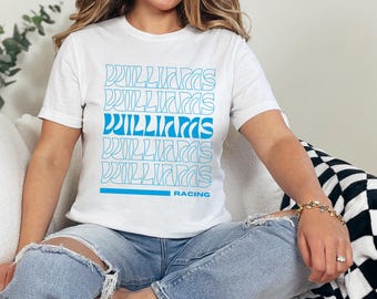 May include: White t-shirt with a blue graphic print of the word "Williams" repeated multiple times in a retro style. The text "Racing" is printed below the graphic.