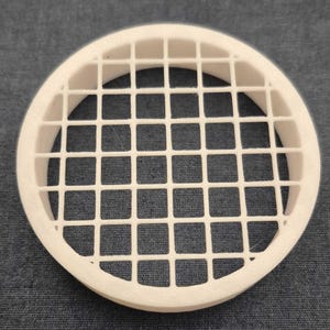 May include: A white ceramic round filter with a grid pattern. The filter has a raised edge and is designed for use in a coffee maker.