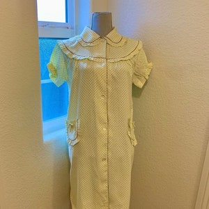 May include: A yellow and brown polka dot dress with a ruffled collar and short sleeves. The dress has a button-up front and two pockets with ruffles. The dress is on a black dress form.