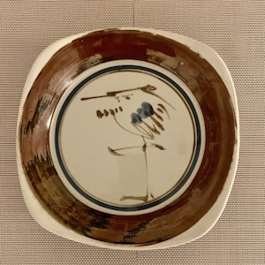 May include: A white ceramic plate with a brown rim and a blue and brown design of a bird on a branch. The plate is on a white placemat.