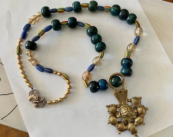 May include: A necklace with a brass pendant and a beaded chain. The chain features dark green, blue, and clear beads with colorful stripes. The pendant is a detailed, ornate brass cross.