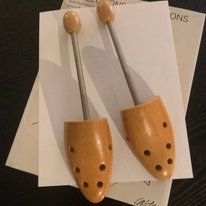 May include: Two wooden shoe trees with metal springs. The shoe trees are light brown and have multiple holes in the wood. The shoe trees are on top of a white piece of paper with the word "Reflections" printed on it.