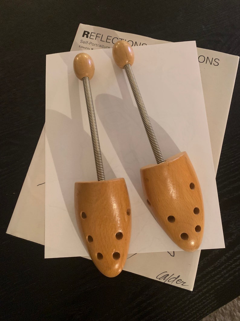 May include: Two wooden shoe trees with metal springs. The shoe trees are light brown and have multiple holes in the wood. The shoe trees are on top of a white piece of paper with the word "Reflections" printed on it.