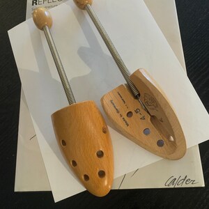 May include: Two wooden shoe trees with metal springs. The shoe trees are made in Germany and have a label that says "4-9". The shoe trees are on a white piece of paper with the word "REFLECTIONS" written on it.