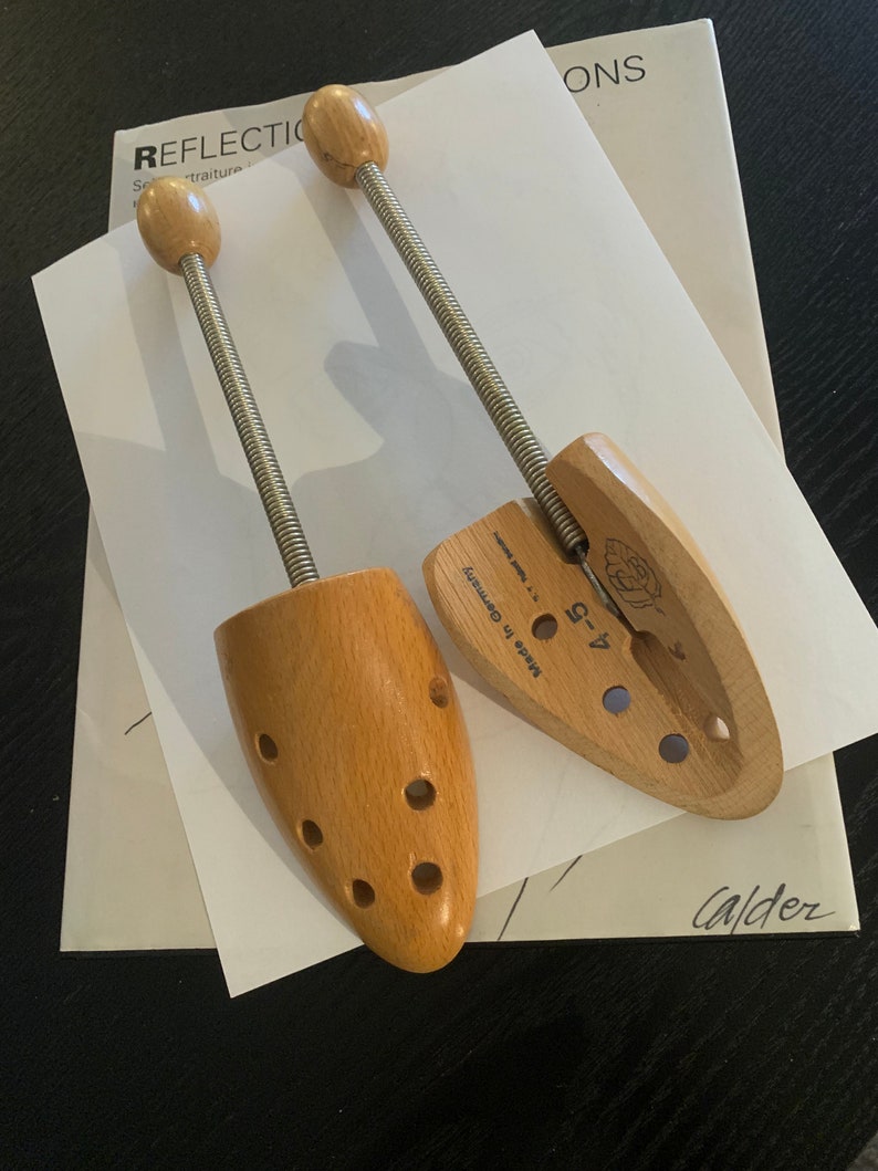 May include: Two wooden shoe trees with metal springs. The shoe trees are made in Germany and have a label that says "4-9". The shoe trees are on a white piece of paper with the word "REFLECTIONS" written on it.