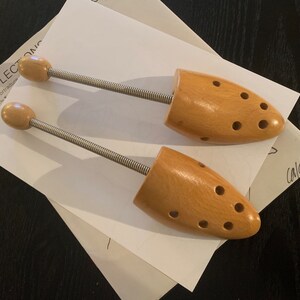 May include: Two wooden shoe trees with metal spring mechanisms. The shoe trees are light brown and have holes for ventilation. The shoe trees are on a white piece of paper with the text "REFLECTONIC Self-Portraits" and "Calder" written on it.