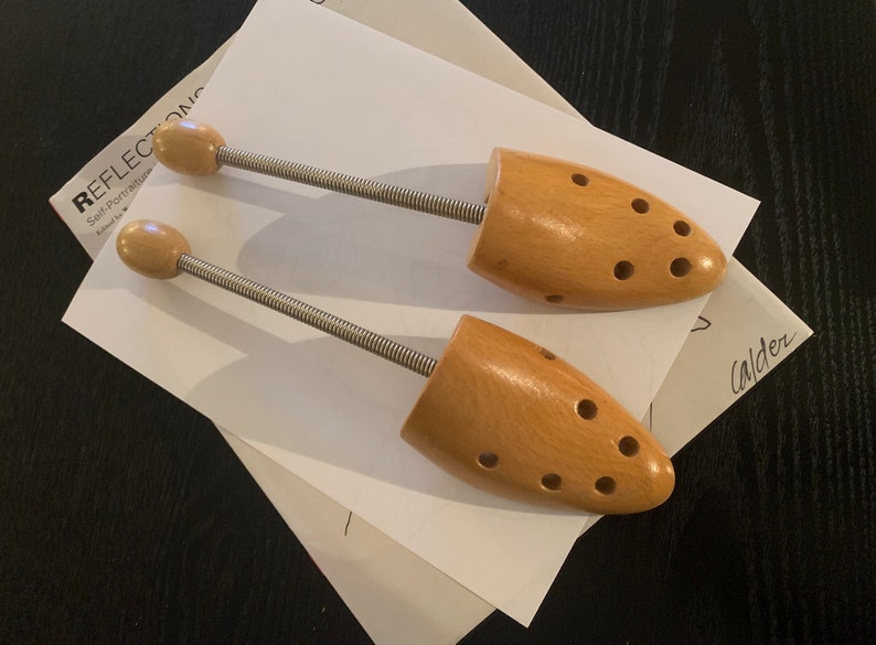 May include: Two wooden shoe trees with metal spring mechanisms. The shoe trees are light brown and have holes for ventilation. The shoe trees are on a white piece of paper with the text "REFLECTONIC Self-Portraits" and "Calder" written on it.