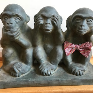 May include: Three gray stone monkeys, each with a different gesture: one covering its eyes, one covering its ears, and one covering its mouth. The monkey covering its ears is wearing a red and blue bow tie.
